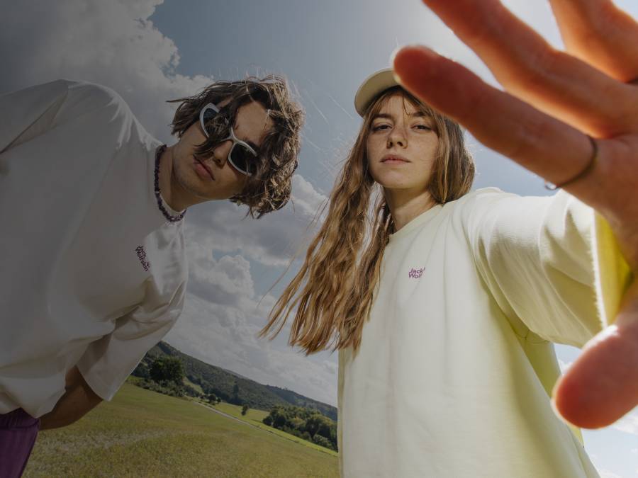 Two young people are standing in a meadow under a cloudy sky. They are wearing light-colored clothing and looking coolly into the&nbsp;camera.